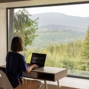 Woman works on laptop while sitting by the table in front of panoramic window with great view on mountains