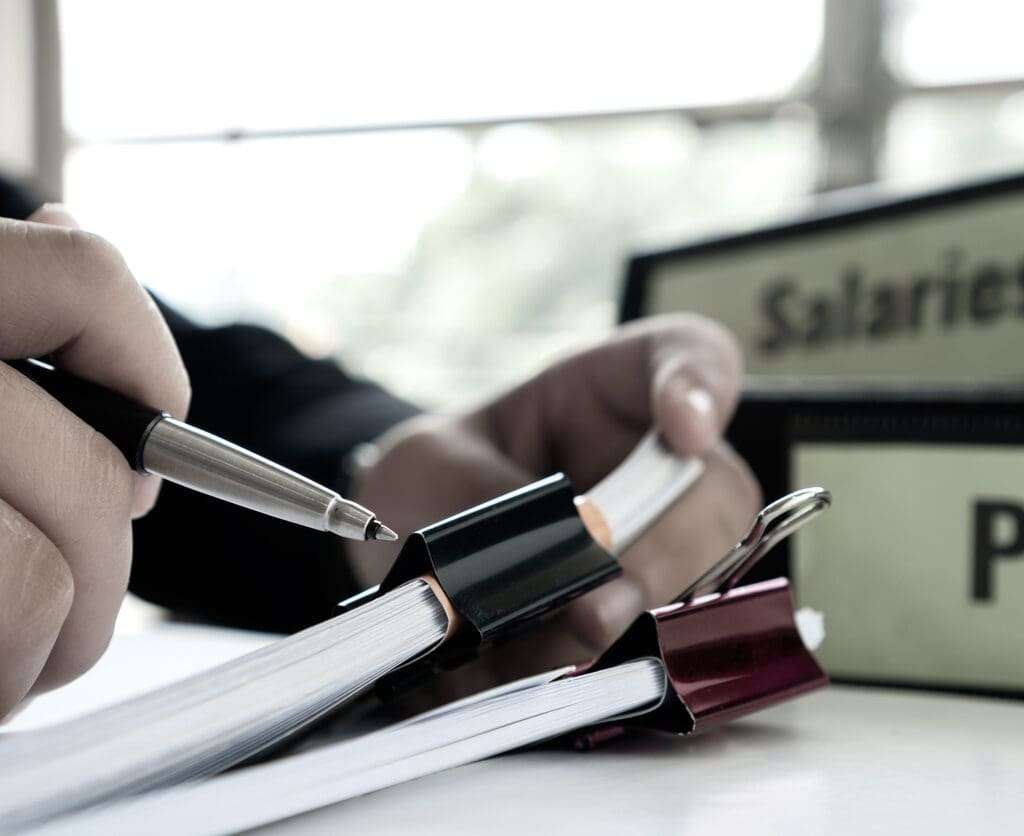 Man holding a pen while signing paperworks near payroll salary binder.