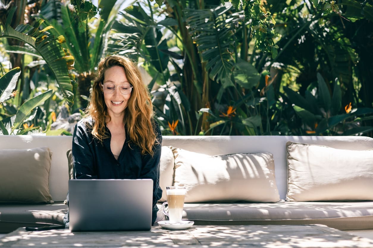 Employee retention strategies - Young woman enjoying remote work in a tropical garden, sitting on a sofa with a laptop and latte.
