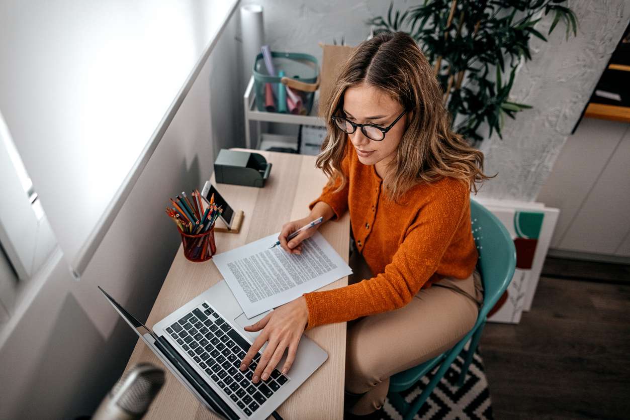 Young woman working on laptop while holding a document at home.