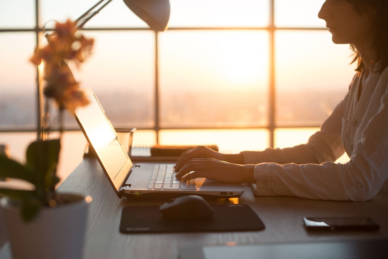 Recognizing remote staffing differences - An unrecognizable female employee working at home with a sunset as a background.