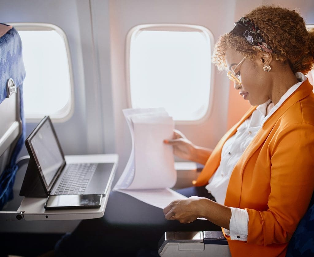 Afro-American businesswoman reading documents and working on digital tablet during flight, depicts use of mobile timekeeping for business trip.