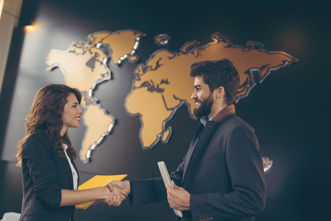 Businesswoman and businessman shake hands in front of a wall with a world map to signify international expansion partnership