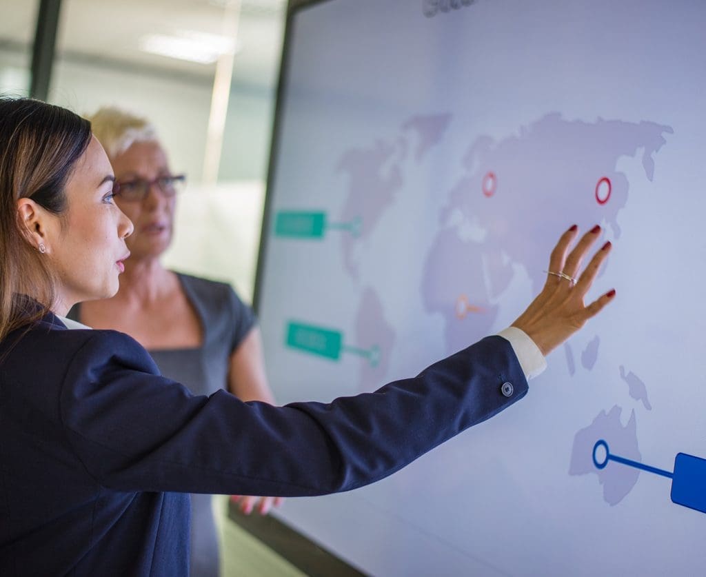 Two businesswomen looking at business targets on a world map on the screen